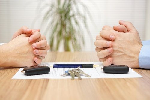 Two pairs of folded hands across from each other, resting on a piece of paper with a pen, two key fobs, and a set of keys on it.